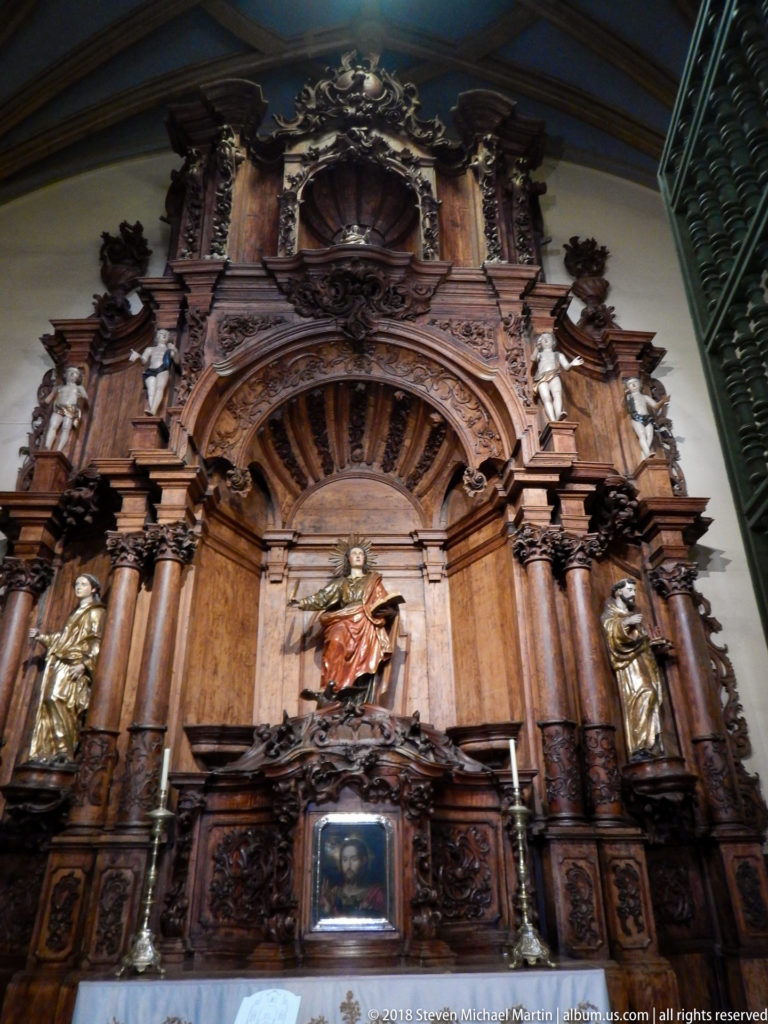 Dark carved wooden retablo with a central saint figure, flanking sculptures, columns, niches, and elaborate upper ornament.