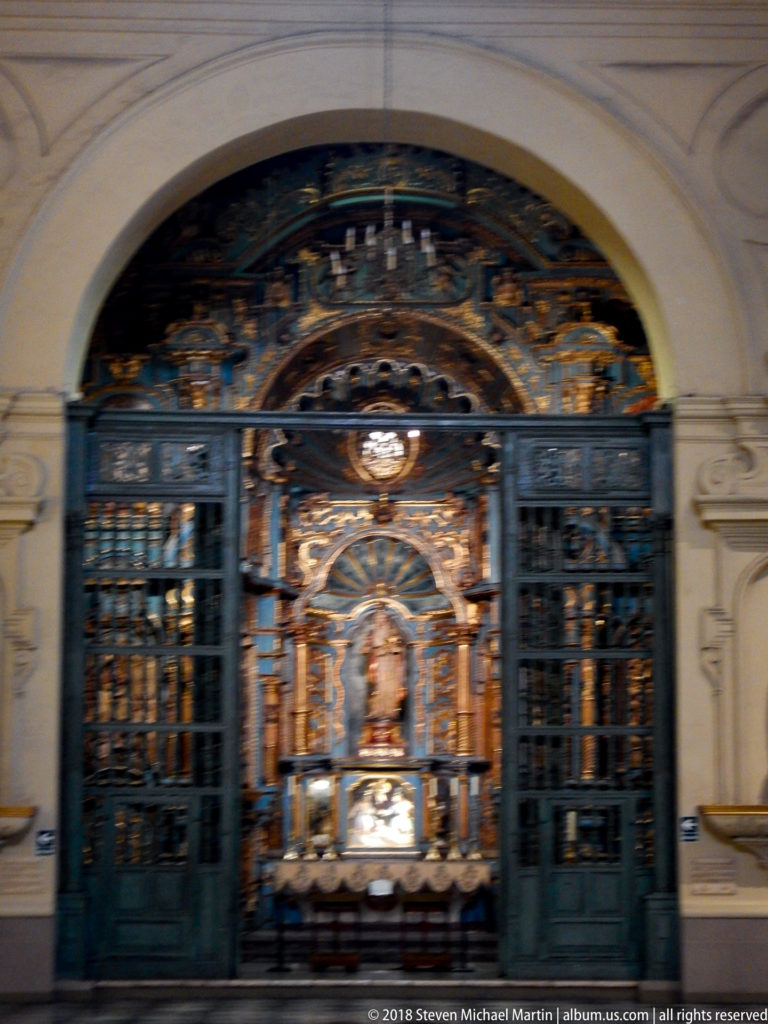 Dark metal gates frame a richly ornamented chapel shrine with gilded details and a small central sacred figure.