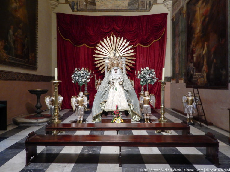 Crowned white-robed Virgin statue before red drapery and radiant halo, flanked by four smaller figures, flowers, and candles.