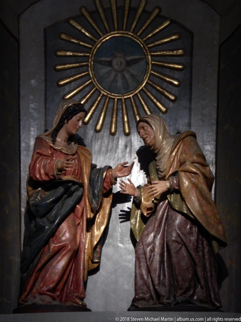 Two polychrome female sacred figures facing each other beneath a radiant circular emblem with a dove.