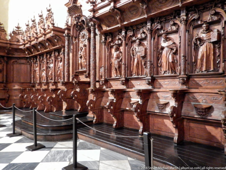 Oblique row of richly carved wooden choir stalls with high backs, standing figures, columns, and projecting seats.