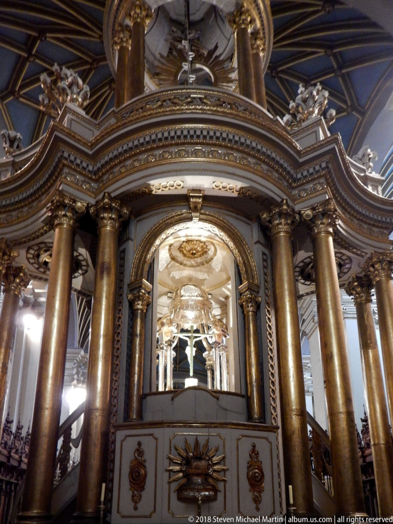 Frontal view of ornate columned canopy with gilded trim, pale paneled base, and bright arched interior.