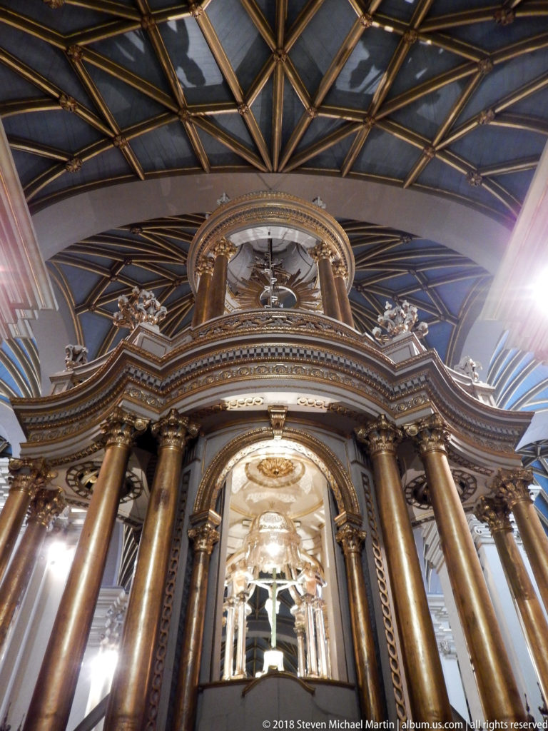 Low-angle view of tall gilded brown columns supporting an ornate canopy beneath a dark geometric ceiling.