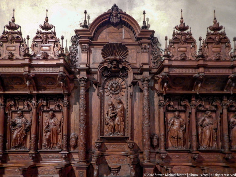 Frontal view of ornate wooden choir stalls with standing carved figures in niches, columns, and a tall central arched bay.