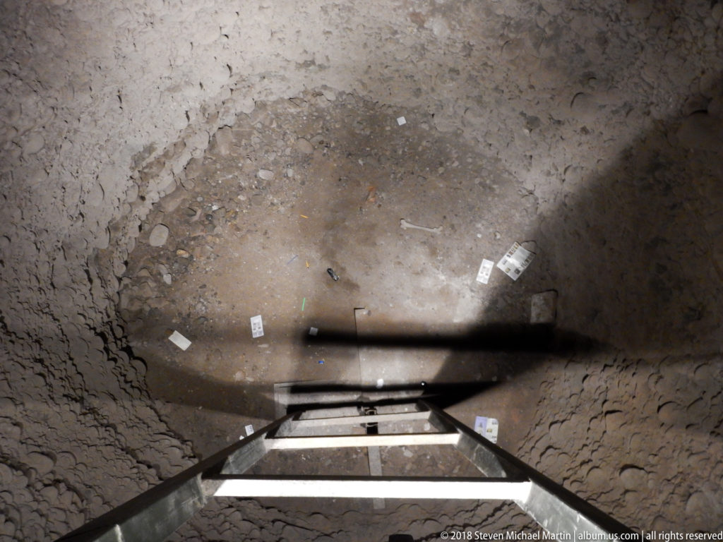 Metal ladder descending into a deep excavation pit with rough walls and small numbered archaeological markers on the floor.