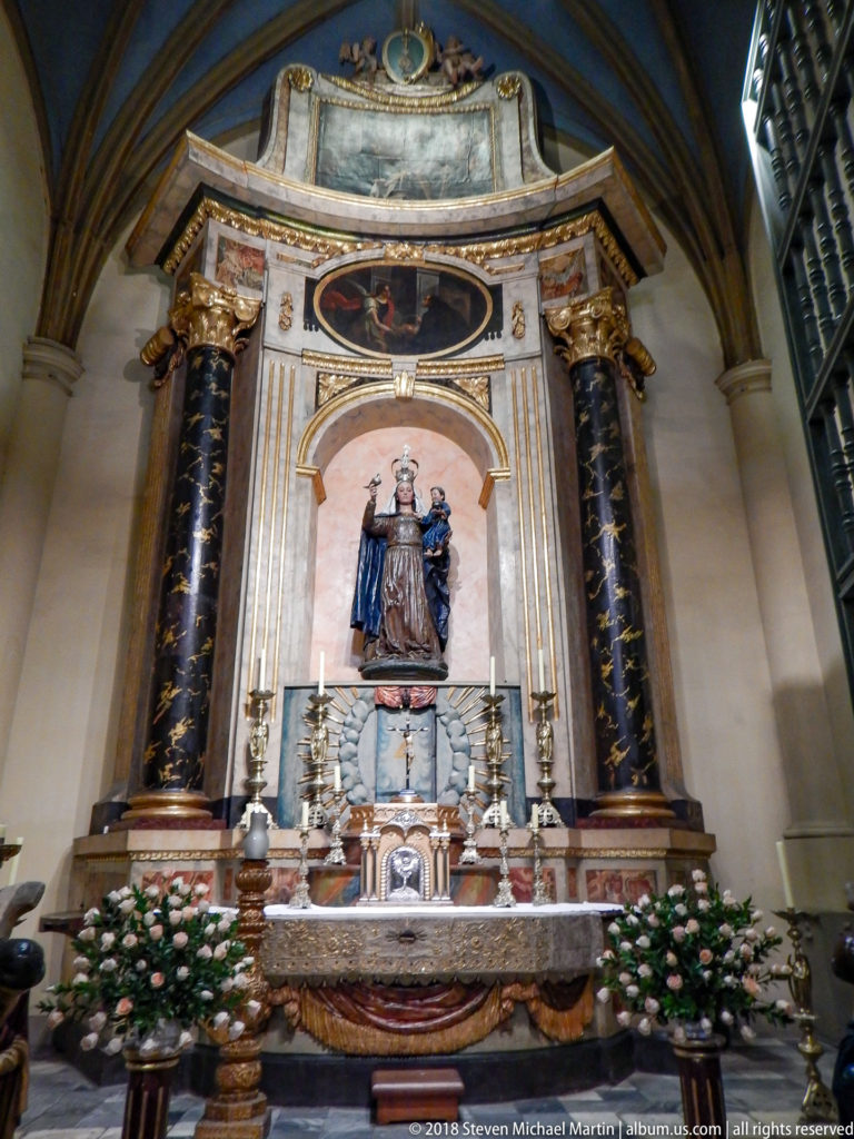 Marble side altar with dark columns, gilded details, flowers, candles, and a central Virgin-and-Child sculpture.
