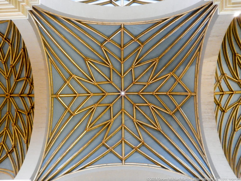 Upward view of a pale cathedral vault crossed by gilded ribs forming stars and geometric polygons.