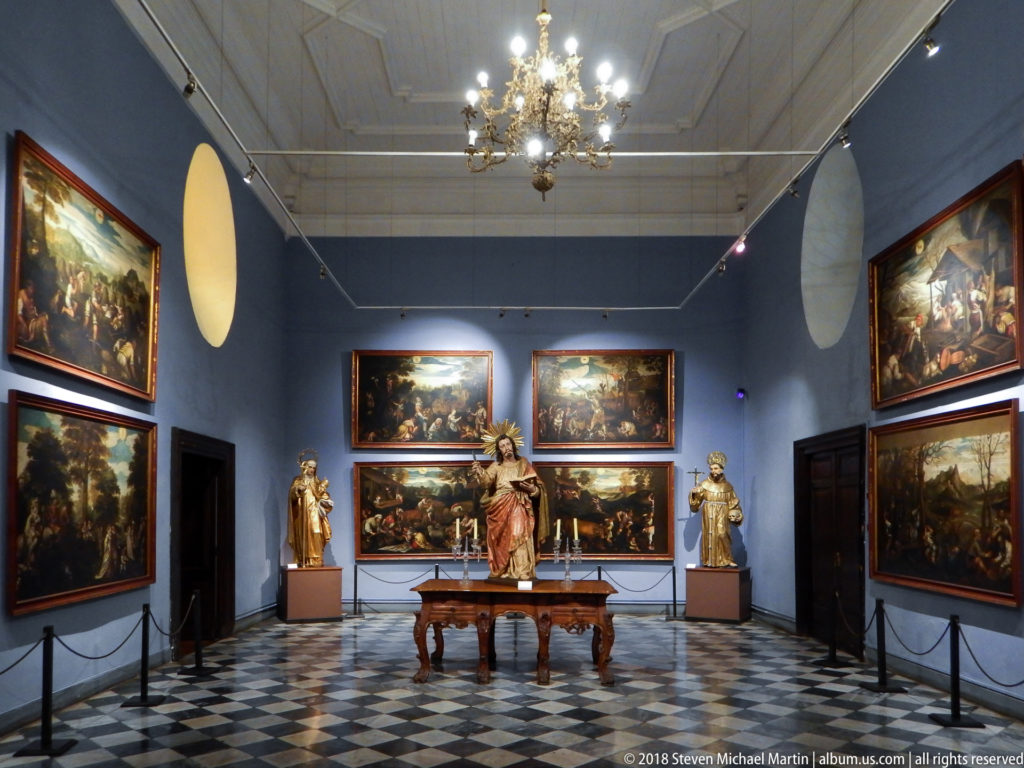 Centered view of blue religious-art gallery with chandelier, patterned floor, paintings, and standing sculptures.