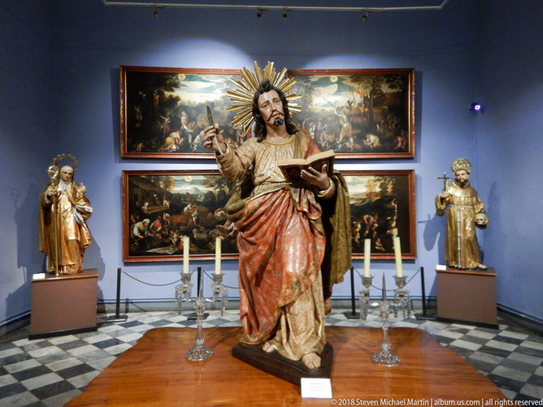 Standing polychrome Christ with radiant halo and open book, flanked by candlesticks, smaller saints, and paintings.