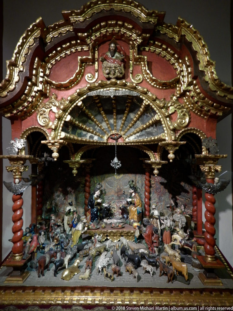 Miniature Nativity with many figures and animals beneath an ornate red-and-gold canopy with twisted columns.