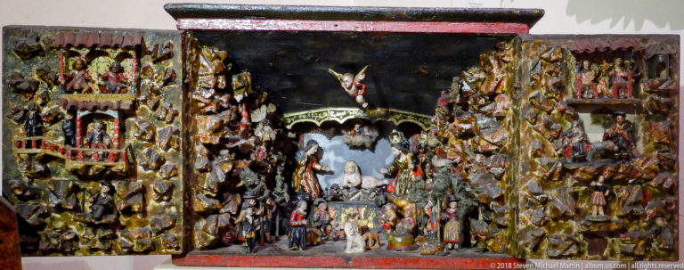 Dense miniature religious diorama with a central Nativity scene, angel, and many figures arranged in side compartments.