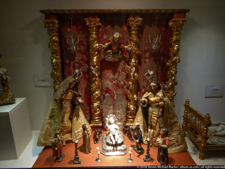 Elaborate miniature Nativity display with Holy Family, many small figures, gilded columns, red backdrop, and flanking statues.