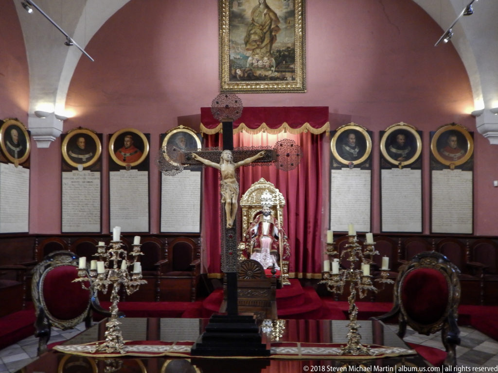 Crucifix above an ornate throne with a seated clerical figure, red drapery, candelabra, chairs, and portrait panels.