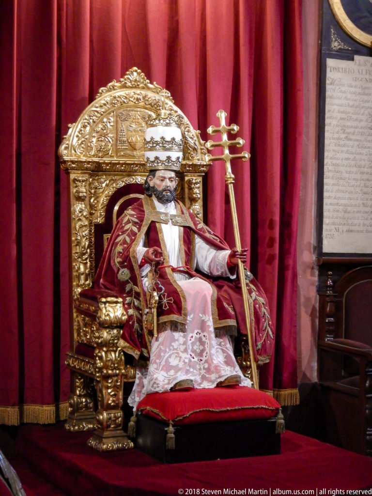 Seated vested clerical figure holding a pastoral staff on an ornate gilded throne before red curtains.