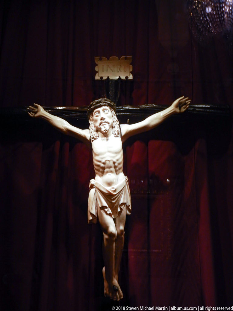 Full crucifix centered against pleated red curtains, with pale sculpted Christ on a dark wooden cross.
