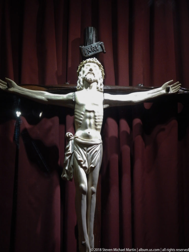 Close frontal view of Christ on a dark cross against deep red drapery, with an inscription panel above.