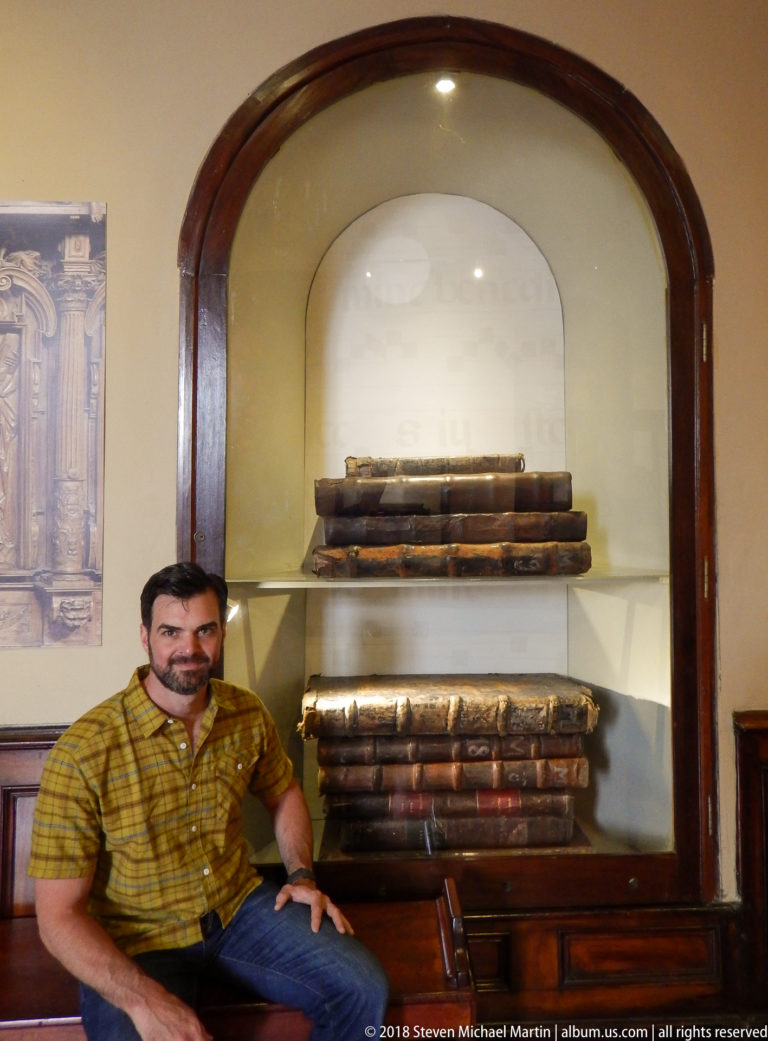 Seated man beside a glass display containing two stacks of oversized, worn leather-bound books.