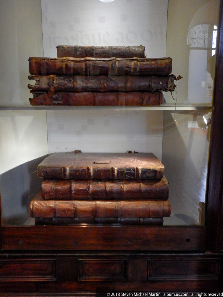 Two stacks of large worn leather-bound volumes displayed behind glass above a dark wooden cabinet.