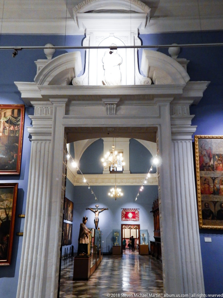 White doorway opening into a blue museum gallery filled with religious paintings, sculptures, chandeliers, and a crucifix.