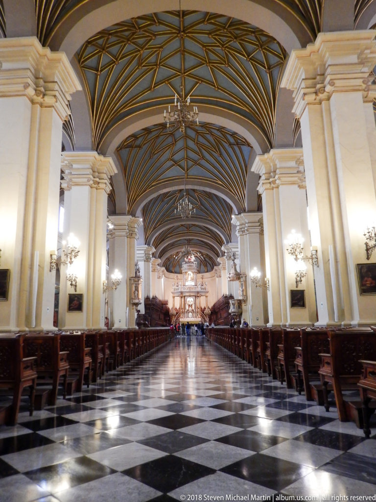 Long central nave with cream piers, wooden pews, checkered floor, and geometric vaulting leading toward the distant sanctuary.