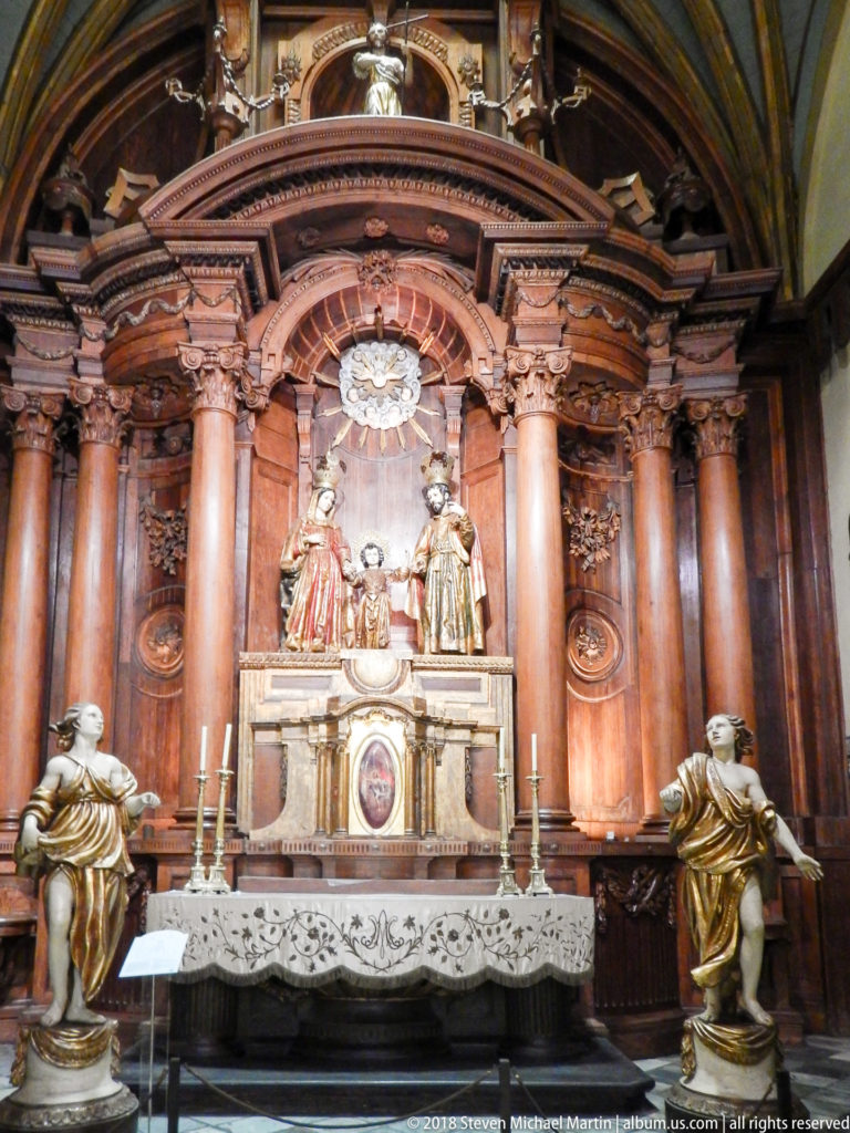 Warm wooden side altar with paired columns, central sculptural group, lace altar cloth, candlesticks, and two flanking statues.