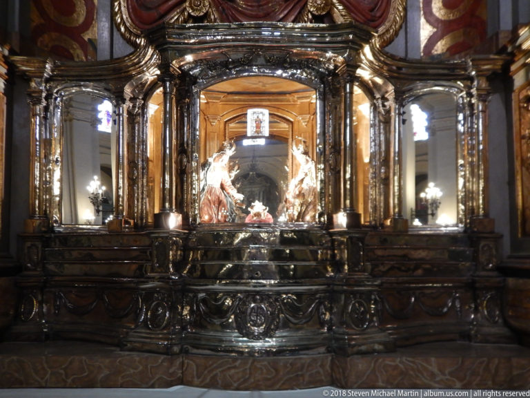 Wide view of a dark glass-fronted shrine with paired columns and a brightly lit sculptural religious group at center.