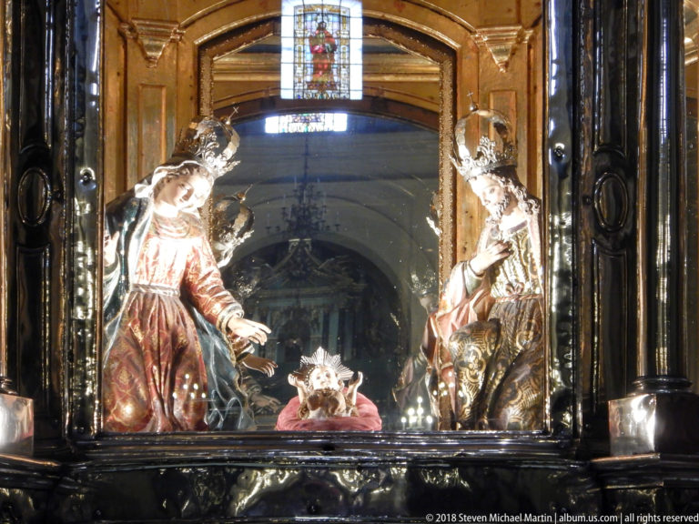 Glass-enclosed polychrome religious tableau with two standing figures leaning toward a smaller central figure in a dark shrine.