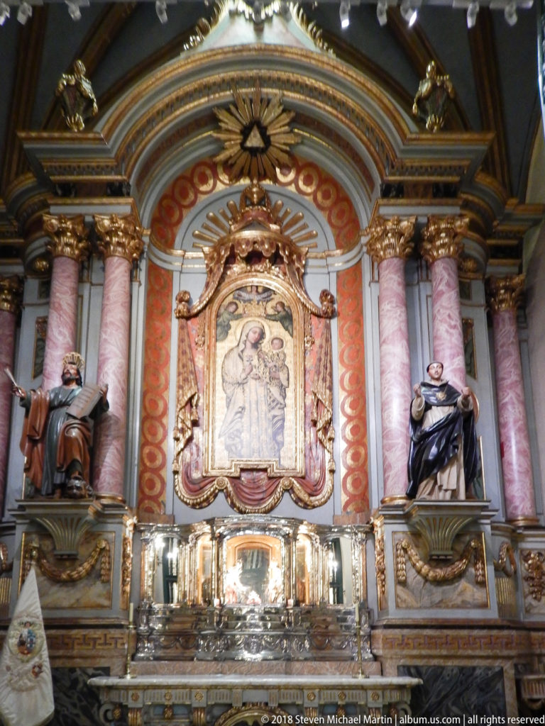 Colorful side altar with pink marbleized columns, central devotional image, seated saint figures, and silver-toned altar objects.