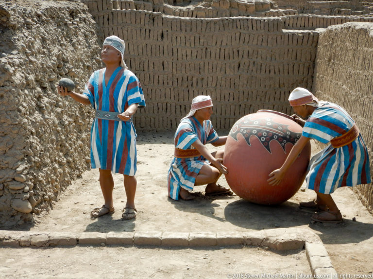 Three costumed interpreters in striped garments beside a large decorated ceramic vessel within reconstructed adobe walls.