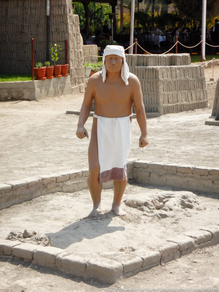 Life-size male interpretive figure in a white loincloth and head covering standing among low adobe walls at Huaca Pucllana.