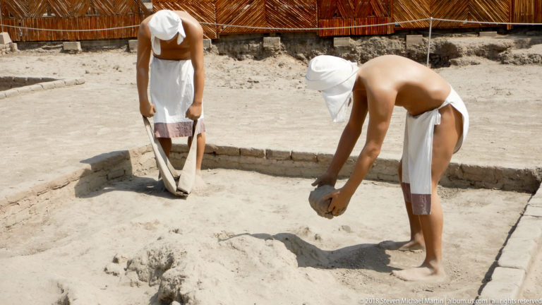 Two life-size reconstructed figures in simple white garments working inside a low adobe enclosure, one bending toward the ground.