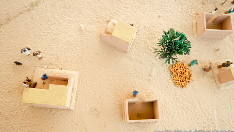 Overhead scale model with small rectangular compounds, miniature people, vegetation, and activity areas on a sandy ground surface.