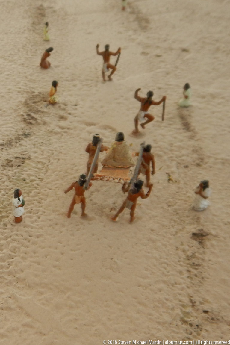Miniature diorama with groups of small figures surrounding and carrying a central platform or burden across sandy ground.