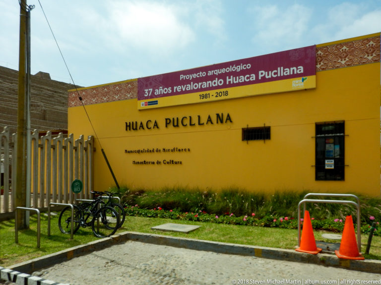 Yellow Huaca Pucllana entrance building with archaeological-project banner, bicycles, flower bed, and traffic cones along the street.