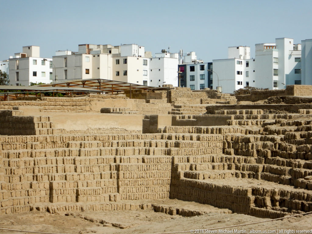 Excavated adobe terraces at Huaca Pucllana with modern white apartment buildings rising directly behind the archaeological site.