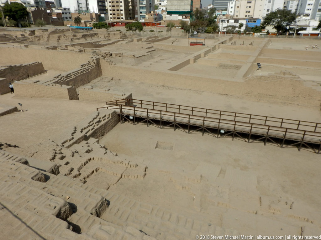 Elevated view over sandy plazas, low excavated walls, and modern walkways at Huaca Pucllana with city buildings beyond.