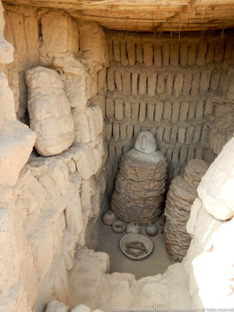 Reconstructed burial chamber with a central wrapped funerary bundle, additional textile-wrapped forms, and ceramic offerings on the floor.