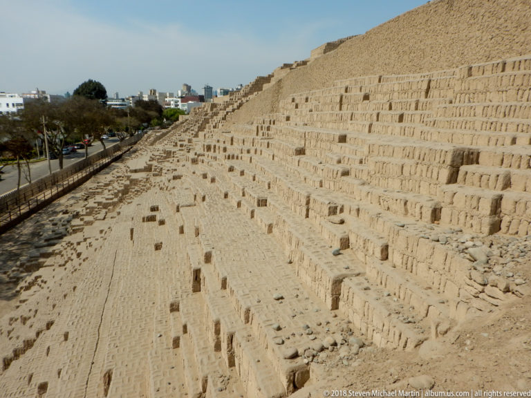 Long sloping adobe wall at Huaca Pucllana beside a fenced, tree-lined urban street with modern buildings in the distance.