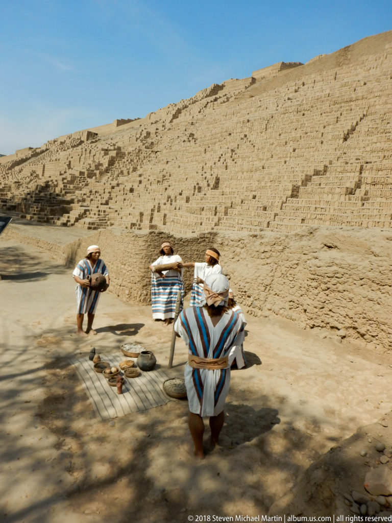 Four people in striped ponchos stand and crouch beside objects at the base of Huaca Pucllana’s tall adobe wall.