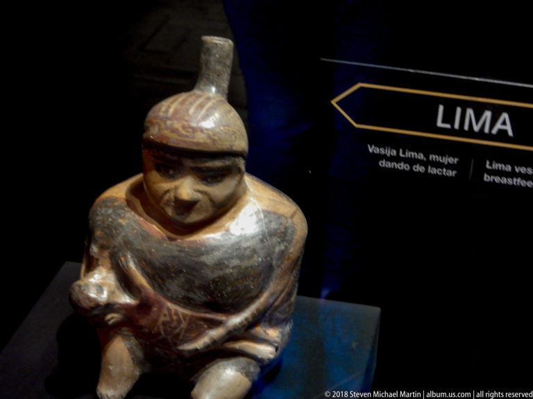 Seated female ceramic figure nursing an infant, displayed beside a museum label identifying a Lima vessel.