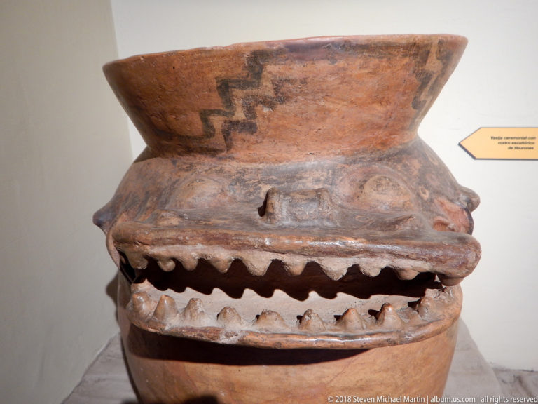 Large reddish-brown effigy vessel with modeled eyes, projecting face, and an open mouth edged by tooth-like forms.