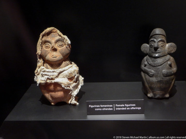 Two female ceramic figurines in a museum case, one pale and wrapped, the other dark and polished, above an offerings label.