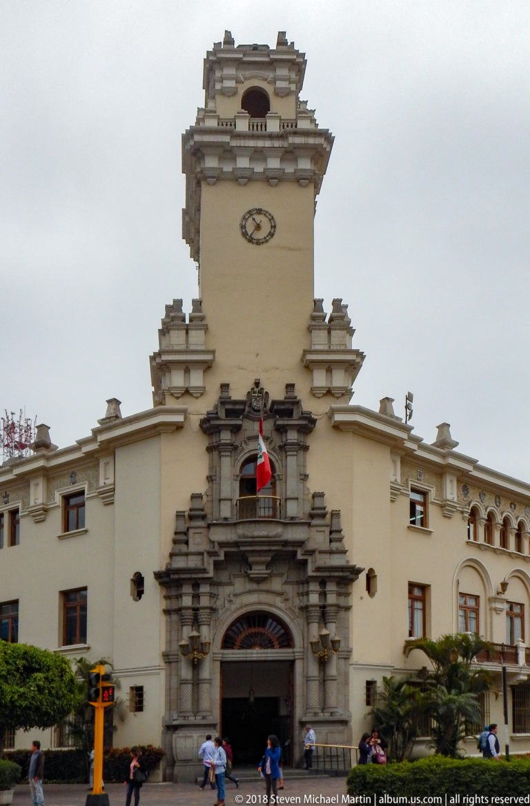 Centered municipal façade with arched entrance, Peruvian flag, dark ornamental stonework, and clock tower rising above.