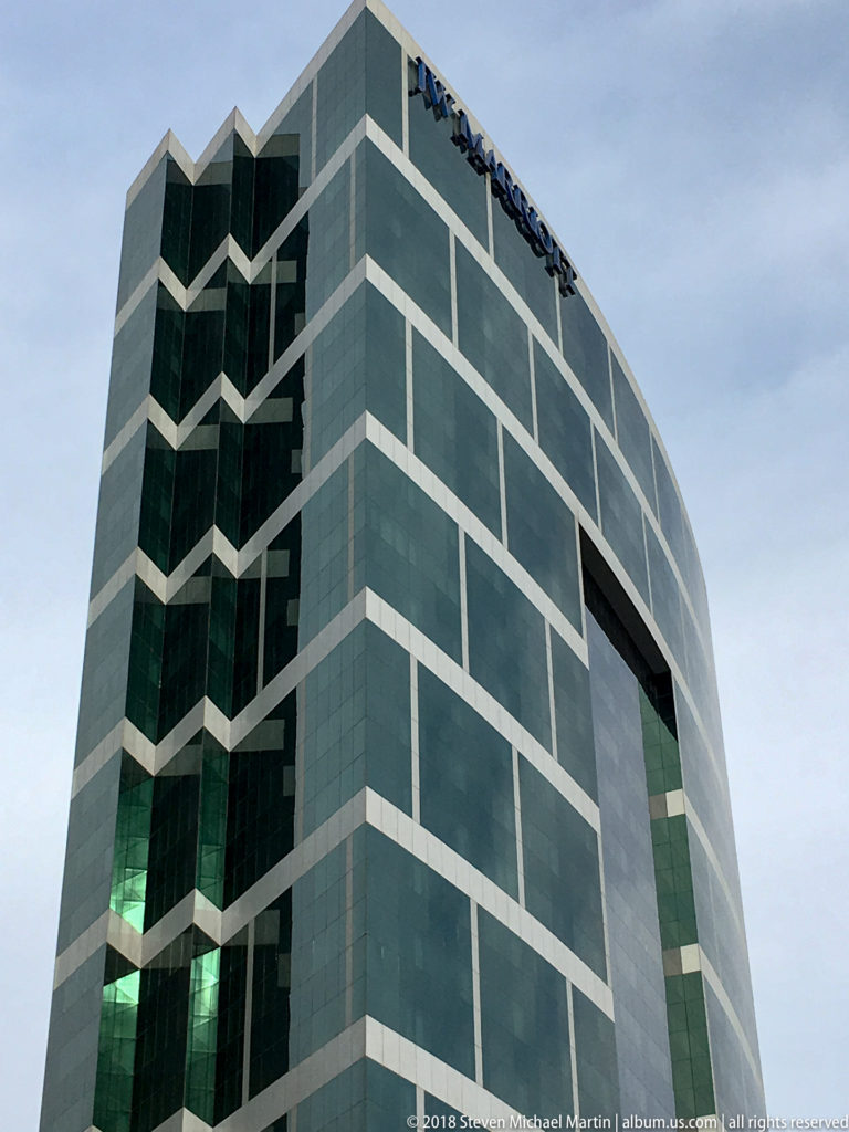 Green-glass high-rise with pale grid bands, a stepped corner profile, and a large dark opening in the façade.