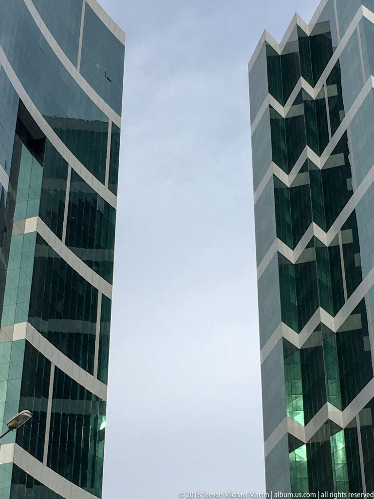 Two dark green glass towers with pale zigzag corner bands rise on either side of a cloudy sky.