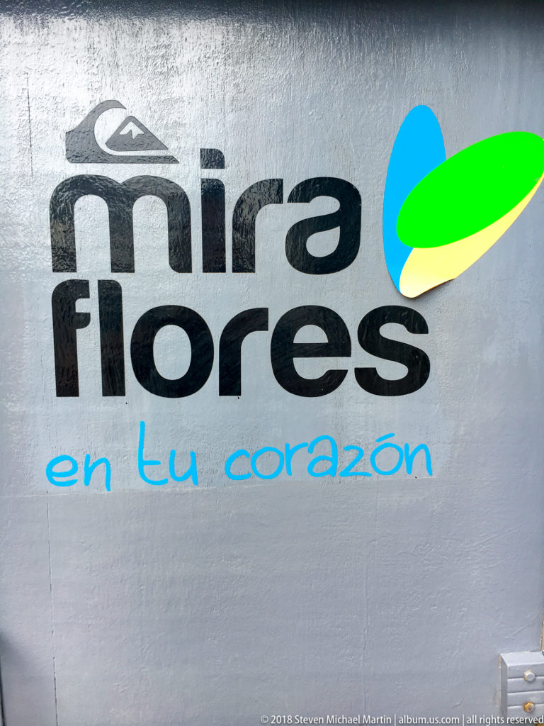 Metallic sign reading “mira flores en tu corazón” with black lettering and a blue, green, and yellow graphic.