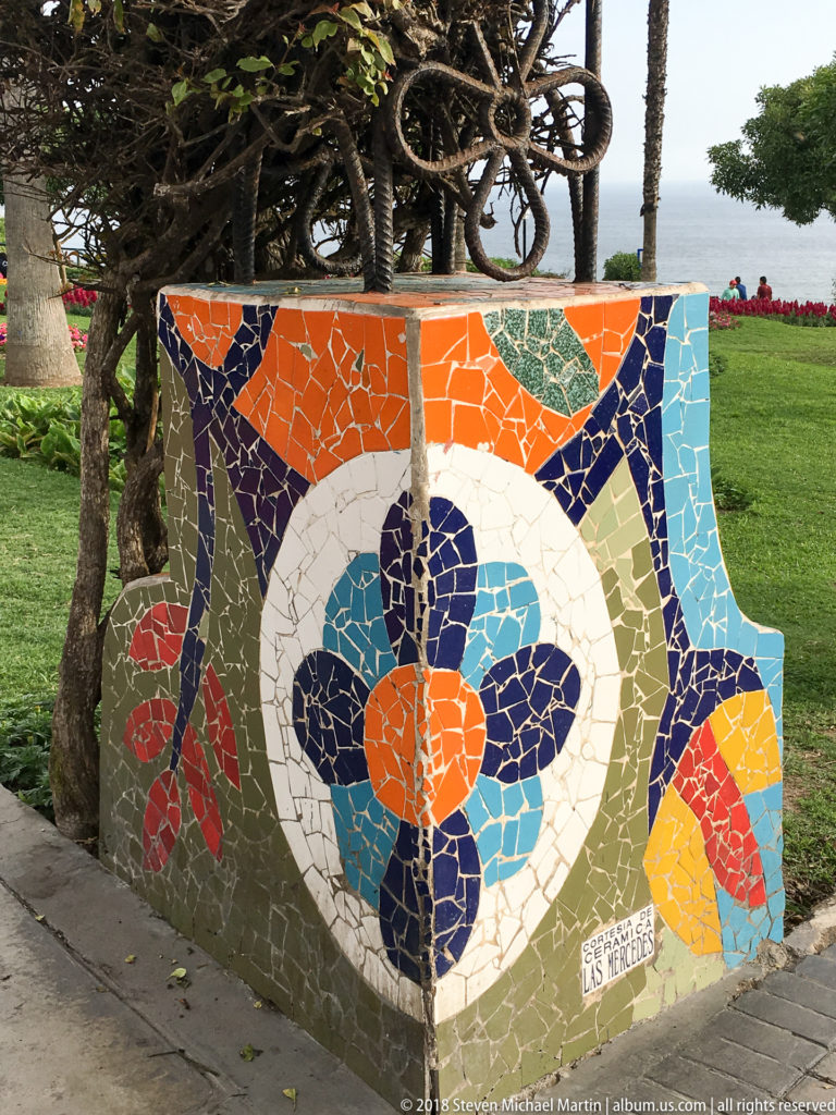 Low park wall covered with a colorful broken-tile floral mosaic, with lawn, trees, and the sea behind it.