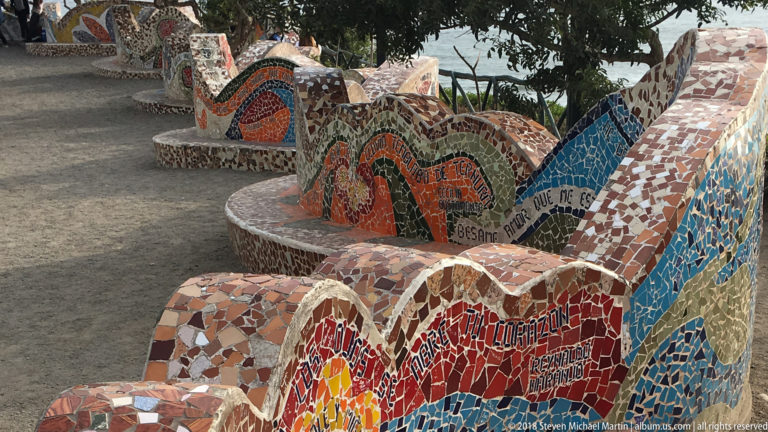Curving park benches covered in multicolored broken-tile mosaics beneath trees, with the ocean visible beyond.
