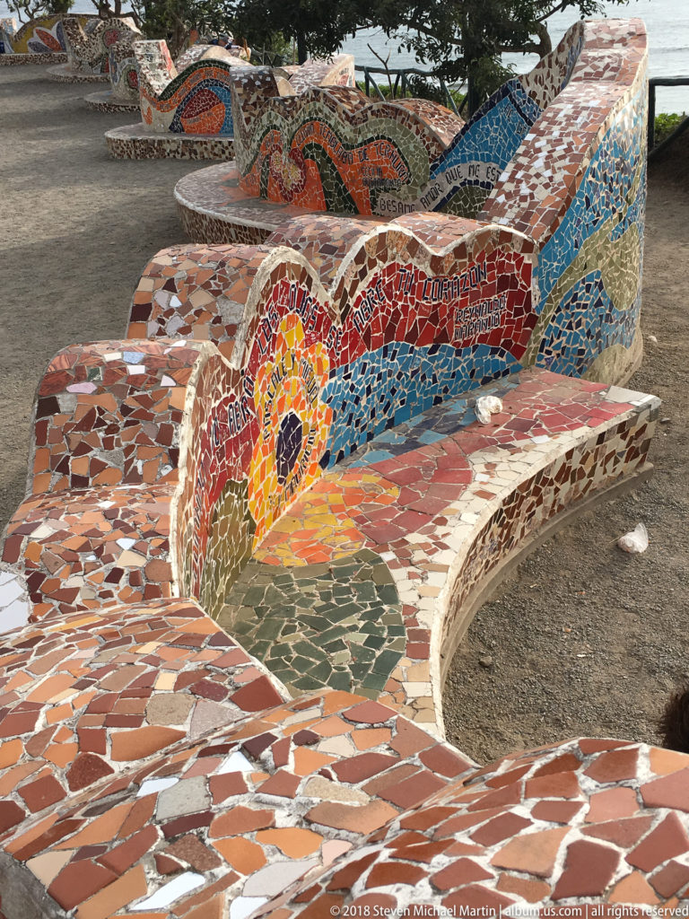 Close oblique view of empty curving mosaic benches with a bright flower motif and red, blue, cream, and brown tile patterns.