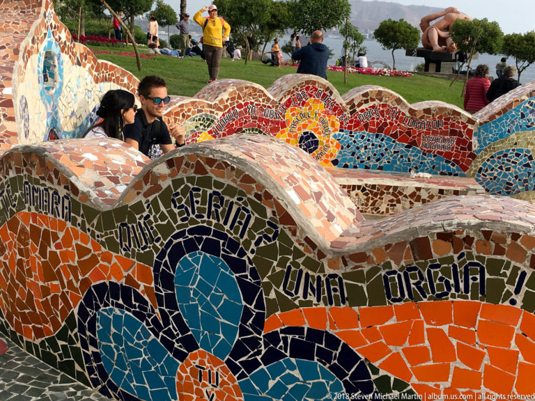 Layered curving mosaic benches with Spanish lettering and colorful motifs at Parque del Amor, with visitors and part of El Beso behind.